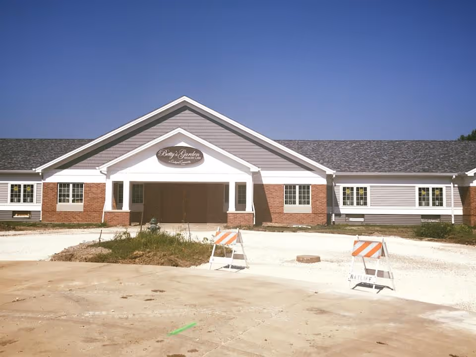 Front exterior view of a single-story building with a gabled roof, brick and siding facade, and a covered entrance. There are two construction barricades in front of the building and some unfinished landscaping. The sky is clear and blue.