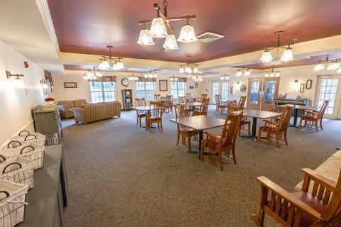 A spacious, well-lit common area with multiple wooden tables and chairs arranged for dining or socializing. The room features carpeted flooring, several chandeliers hanging from the ceiling, and large windows allowing natural light to enter. There are couches and armchairs along the walls, decorative wall clocks, framed pictures, and a sideboard with baskets on the left side.