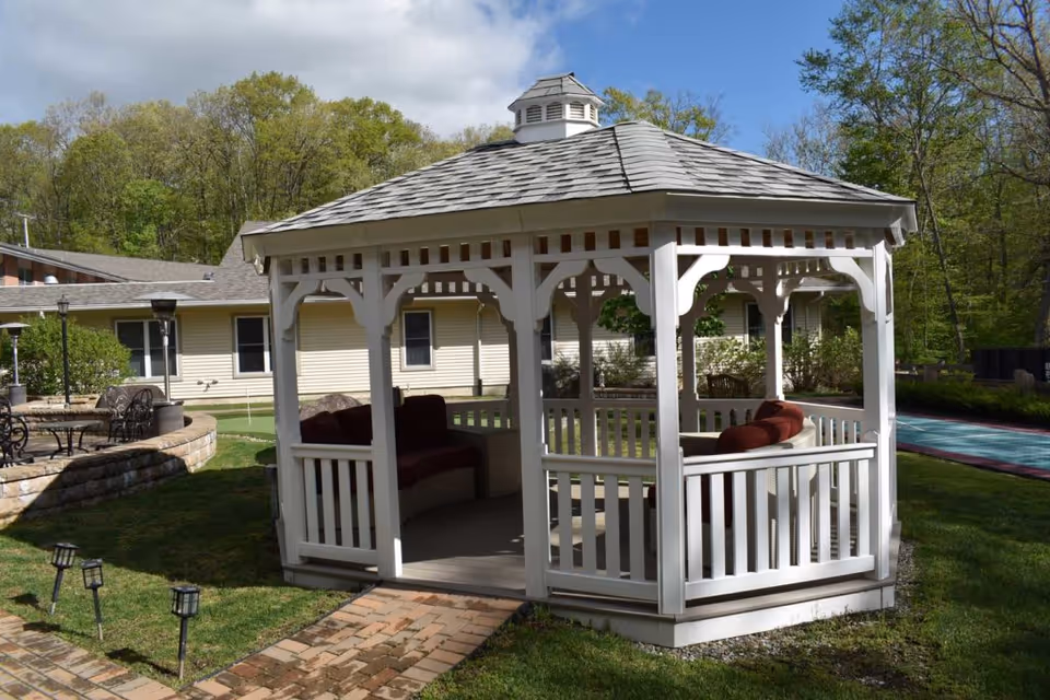 A white wooden gazebo with a shingled roof and cushioned seating inside, situated on a grassy area with a brick pathway leading to it. In the background, there is a beige building with windows and some outdoor furniture, surrounded by trees and greenery under a partly cloudy sky.