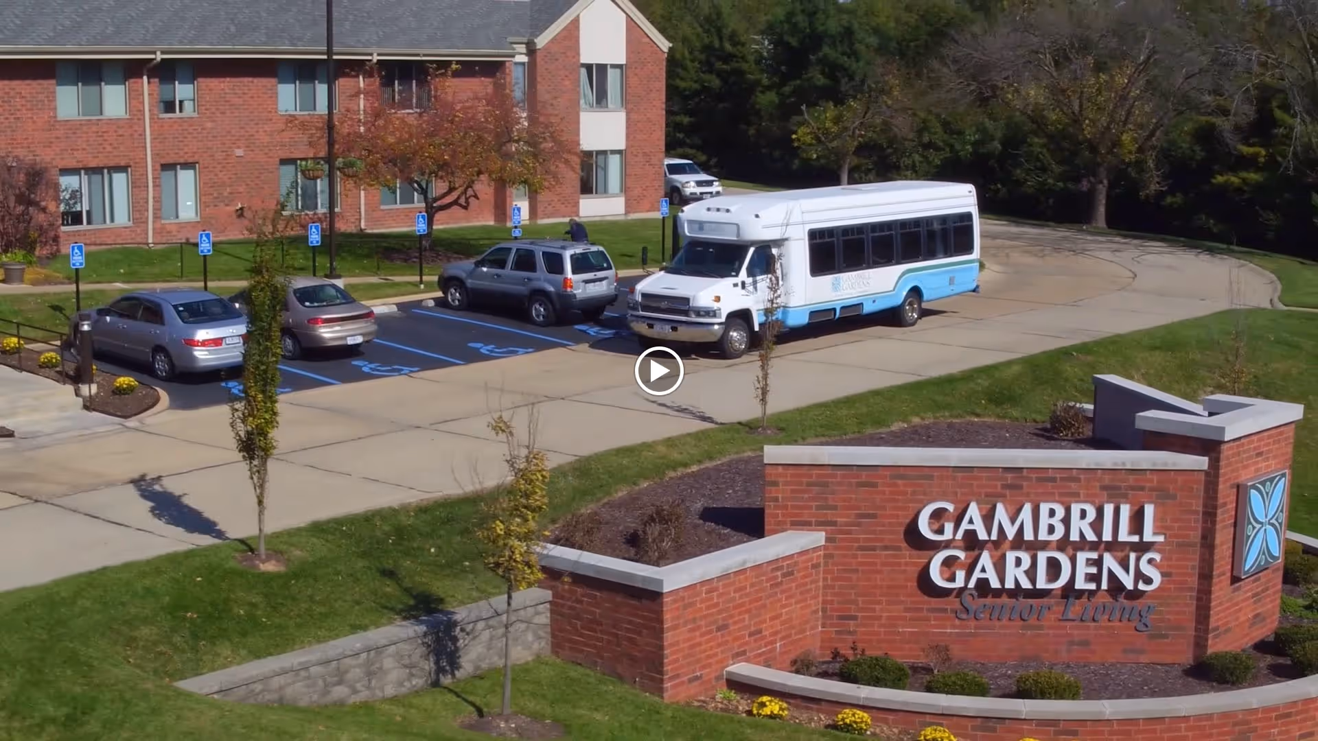 Exterior view of Gambrill Gardens Senior Apartments showing a brick sign with the facility name, a parking lot with several cars and a white and blue shuttle bus, and a two-story brick building in the background surrounded by trees and greenery.
