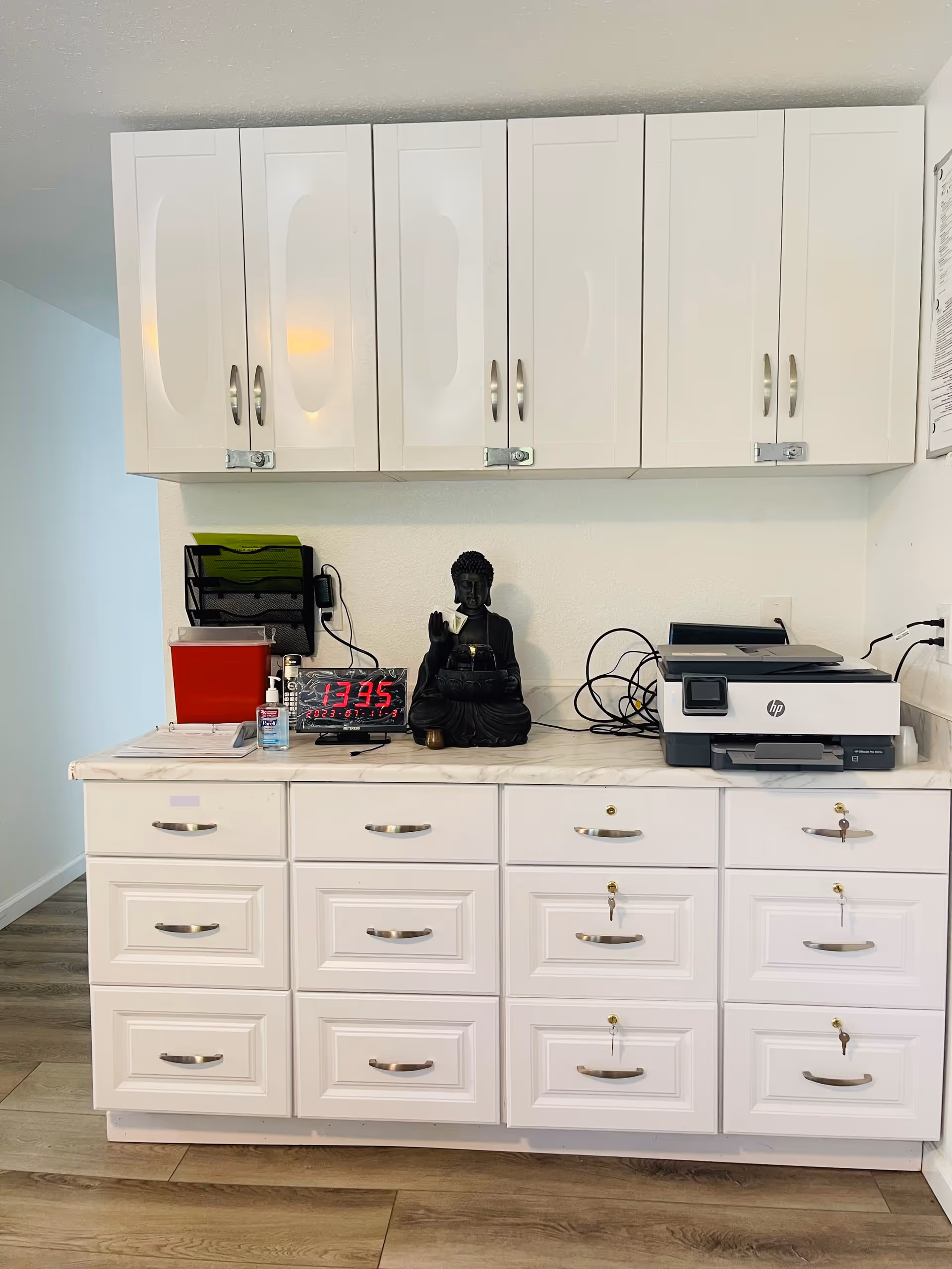 White cabinets with multiple drawers and cupboards, some locked with keys. On the countertop, there is a black Buddha statue, a digital clock displaying the time 13:35, a bottle of hand sanitizer, a red container, a black file organizer with green folders, and an HP printer with cables. The floor is wooden and the walls are white.