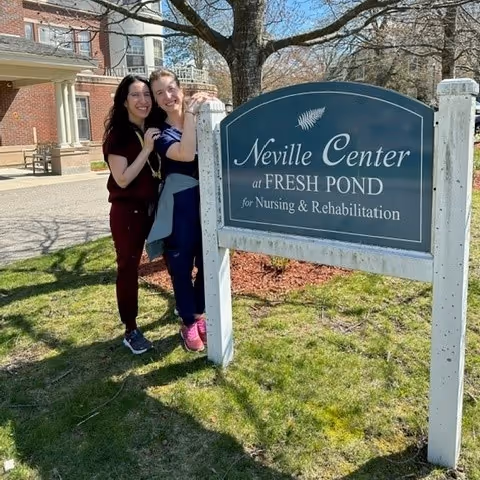 Two women standing outdoors next to a sign that reads 'Neville Center at Fresh Pond for Nursing & Rehabilitation' with a building and trees in the background.