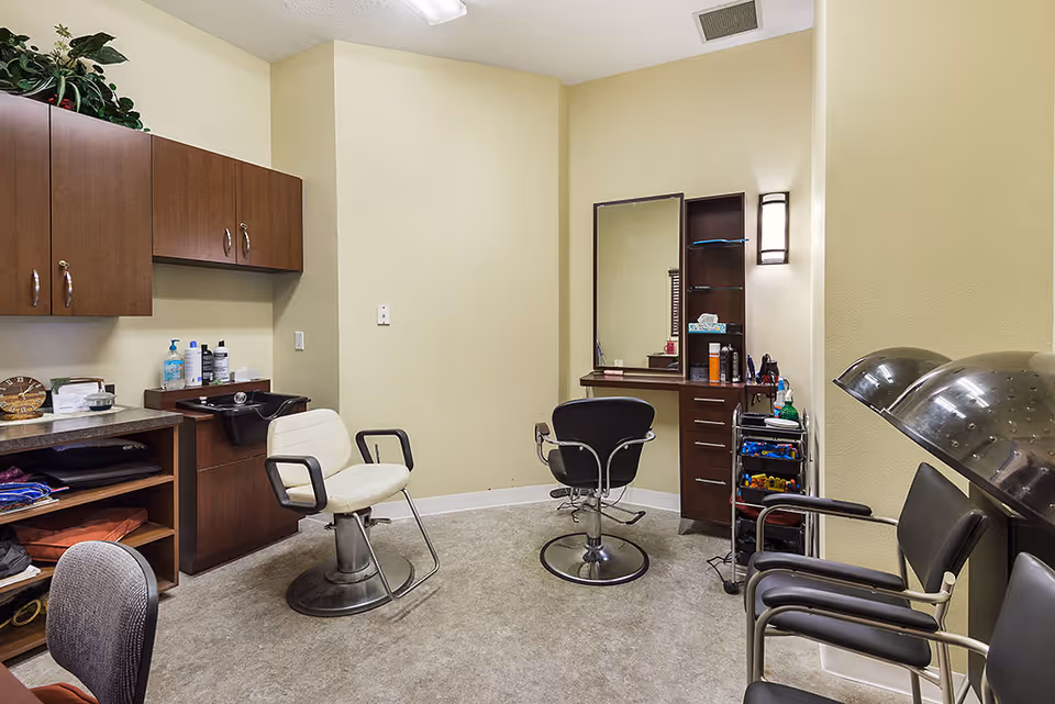 Interior view of a hair salon area in an assisted living facility with two salon chairs, a large mirror, wooden cabinets, hair care products, and two hair dryers with chairs along the right wall.