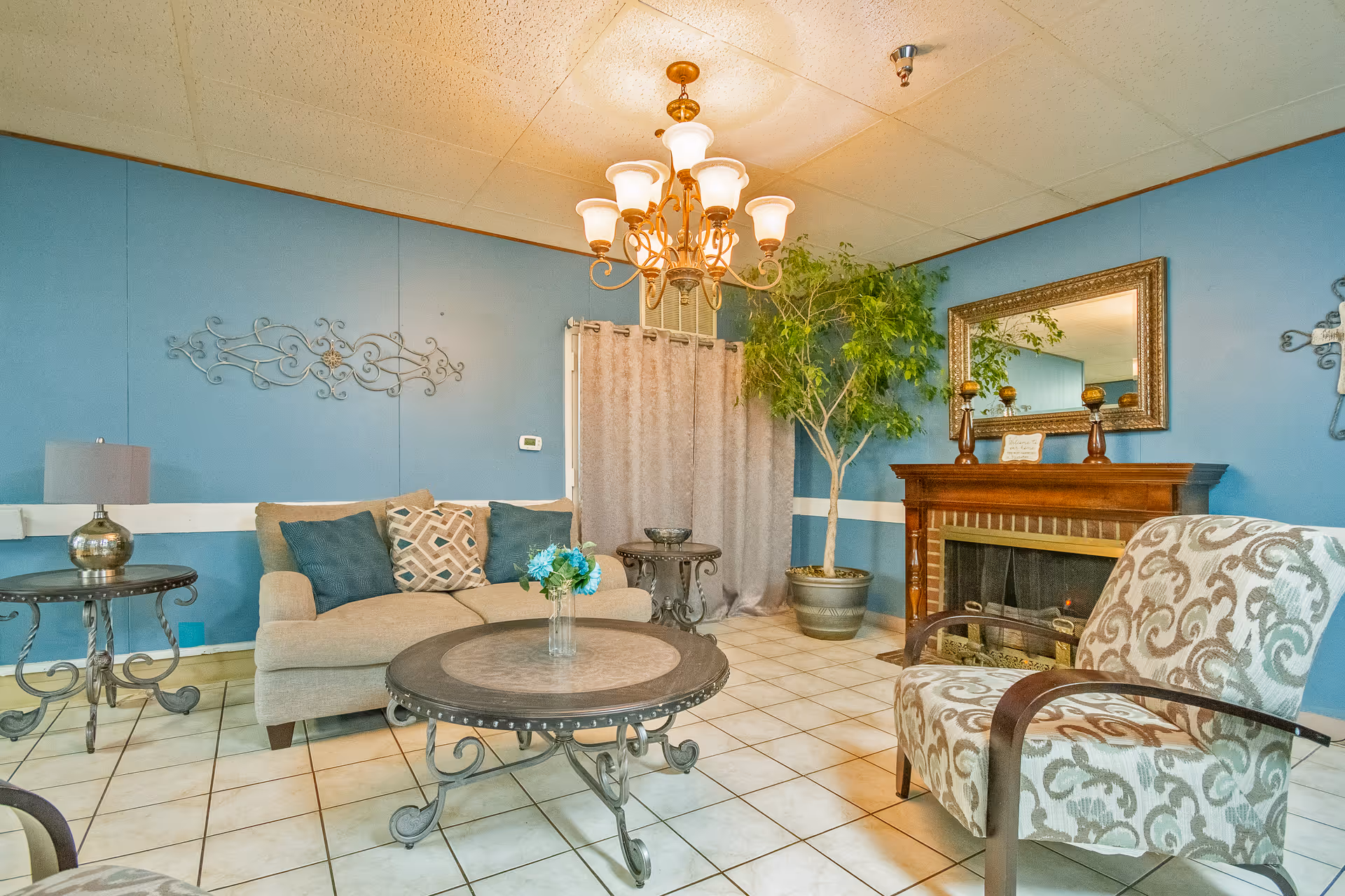 A cozy living room with blue walls and white tiled floor featuring a beige sofa with patterned and solid blue cushions, a round metal coffee table with a small flower vase, a patterned armchair, a wooden fireplace with a large framed mirror above it, a tall potted plant, two side tables with lamps, decorative wall art, and a chandelier hanging from the ceiling.