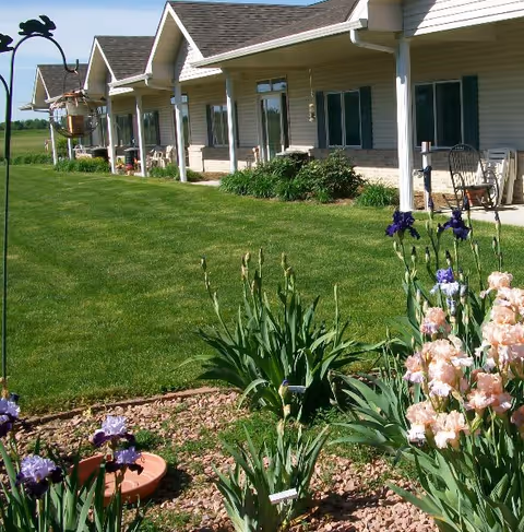 A well-maintained garden area with blooming flowers and green grass in front of a single-story building with a covered porch and several windows. The building has beige siding and a shingled roof, and there are chairs placed on the porch.