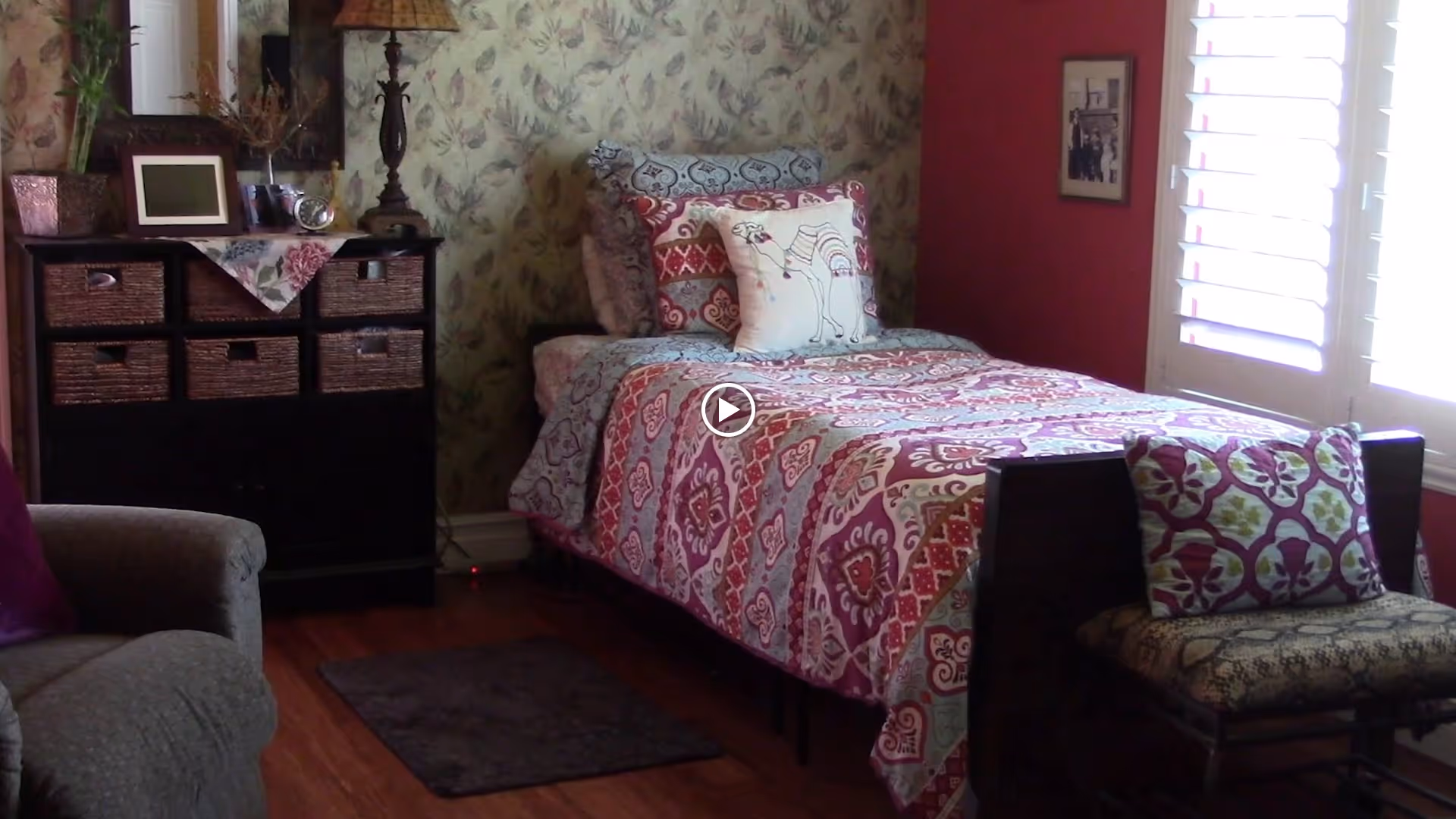 A cozy bedroom with a single bed covered in a patterned quilt and decorative pillows, including one with a camel design. Next to the bed is a cushioned bench with a floral pillow. A dark wooden dresser with woven baskets and decorative items sits against a floral wallpapered wall. A window with white shutters lets in natural light, and a framed picture hangs on the adjacent red wall. A gray armchair is partially visible in the foreground.