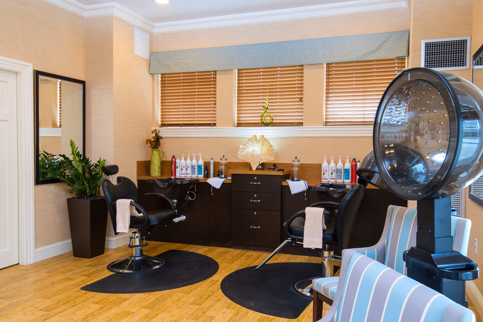 Interior view of a salon area with two black salon chairs in front of a counter with various hair care products. There is a large hair dryer on the right side and a tall plant in a pot on the left. The room has wooden flooring, beige walls, and windows with wooden blinds.