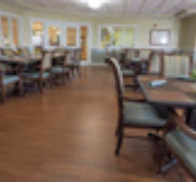 A dining room with multiple wooden tables and chairs arranged neatly on a wooden floor. The room has large windows and a bright, welcoming atmosphere.