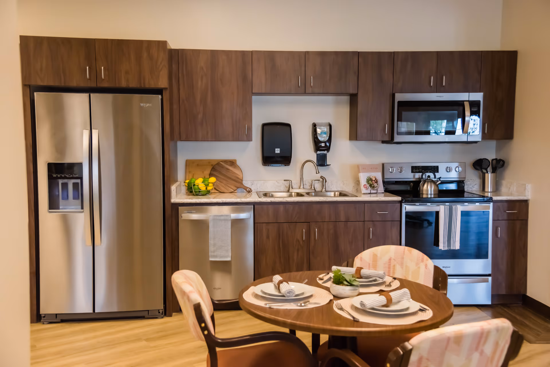 Modern kitchen area with stainless steel refrigerator, dishwasher, stove, and microwave. Wooden cabinets and a round dining table set with plates, napkins, and utensils are visible. The kitchen has a clean and organized appearance.