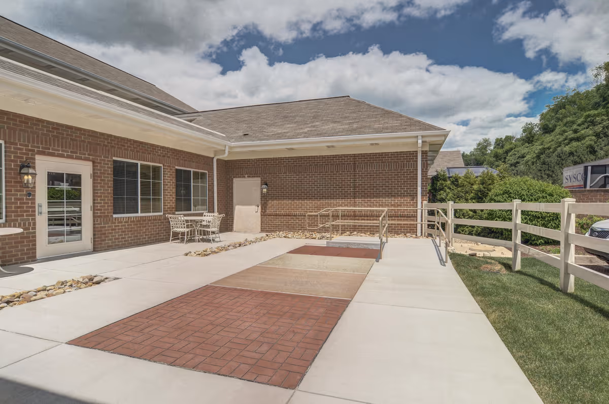 Brick senior living facility courtyard with accessible ramp, outdoor seating, and walkways under a partly cloudy sky.
