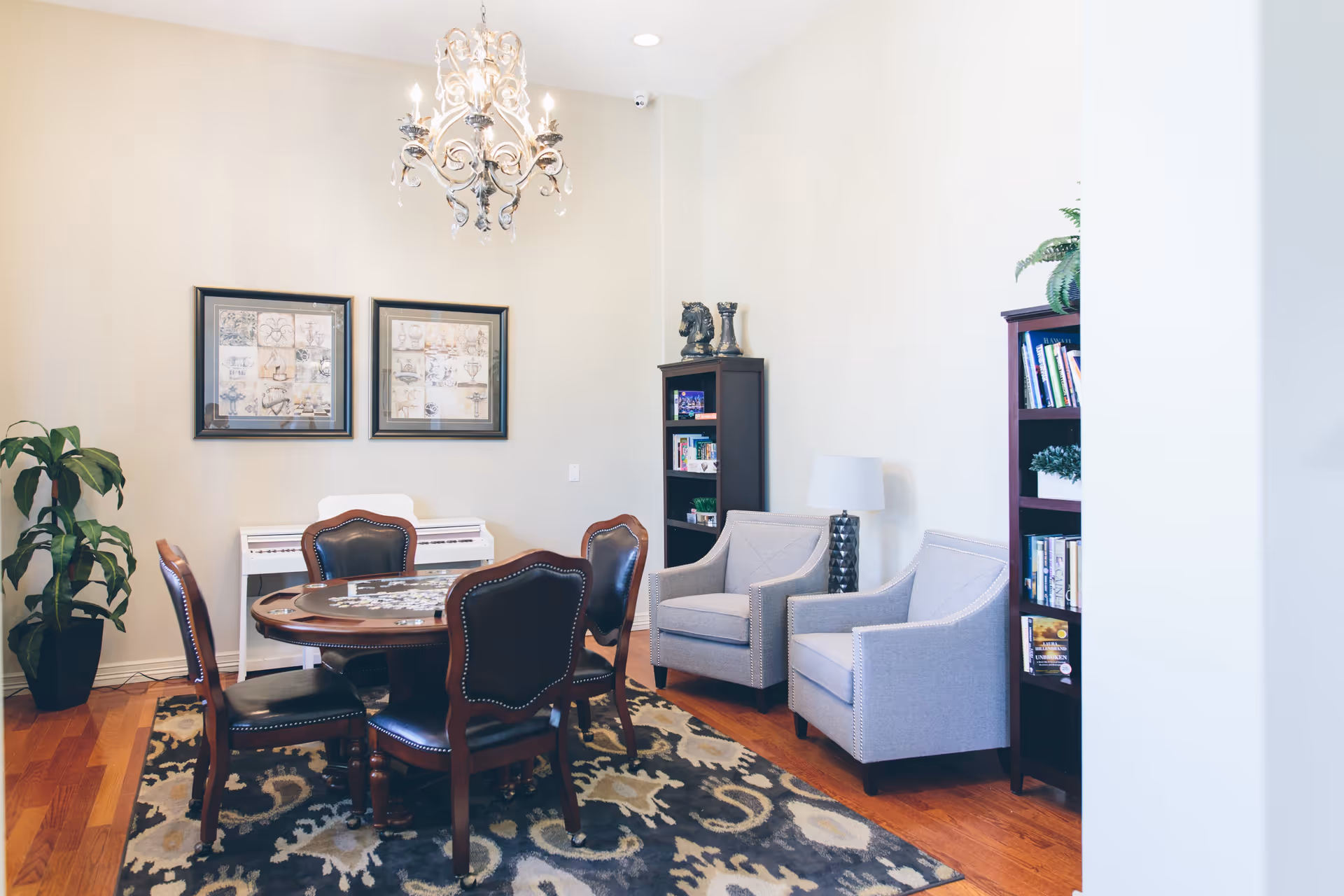A cozy room with a round wooden table surrounded by four dark leather chairs on a patterned rug. Two light gray armchairs are positioned near a tall bookshelf filled with books and decorative items. A white piano is against the wall beneath two framed artworks. A chandelier hangs from the ceiling, and a potted plant is in the corner.