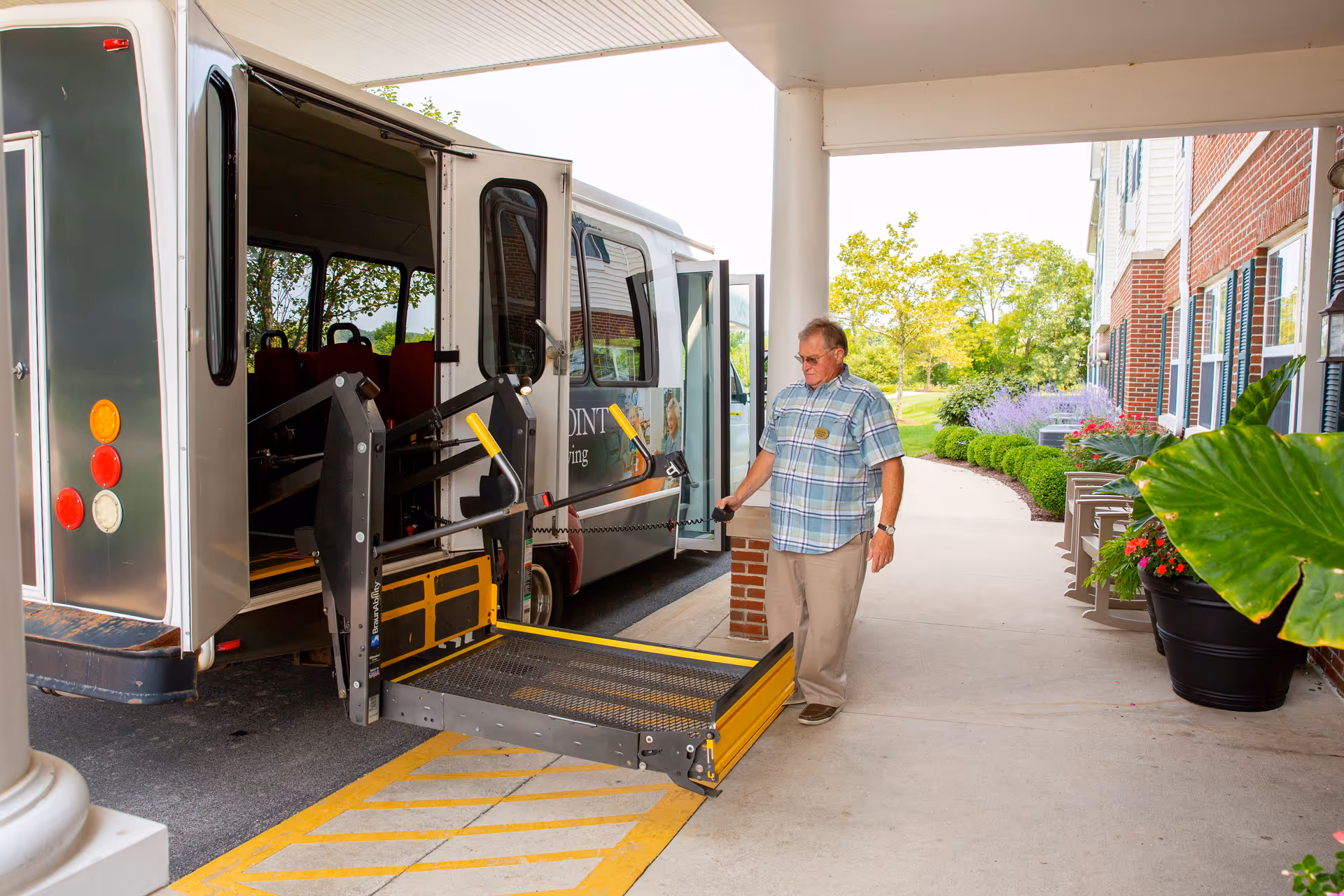 An elderly man standing next to a van with an extended wheelchair lift ramp outside a building entrance with plants and greenery nearby.