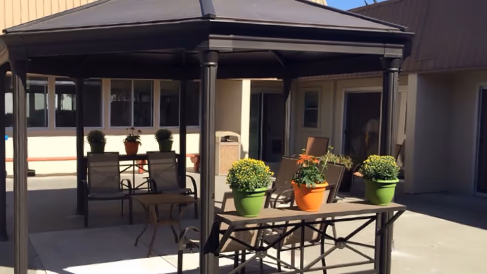 Outdoor patio area at Crestwood Manor featuring a gazebo with seating underneath, several potted plants with flowers on tables, and surrounding building walls with windows and doors.