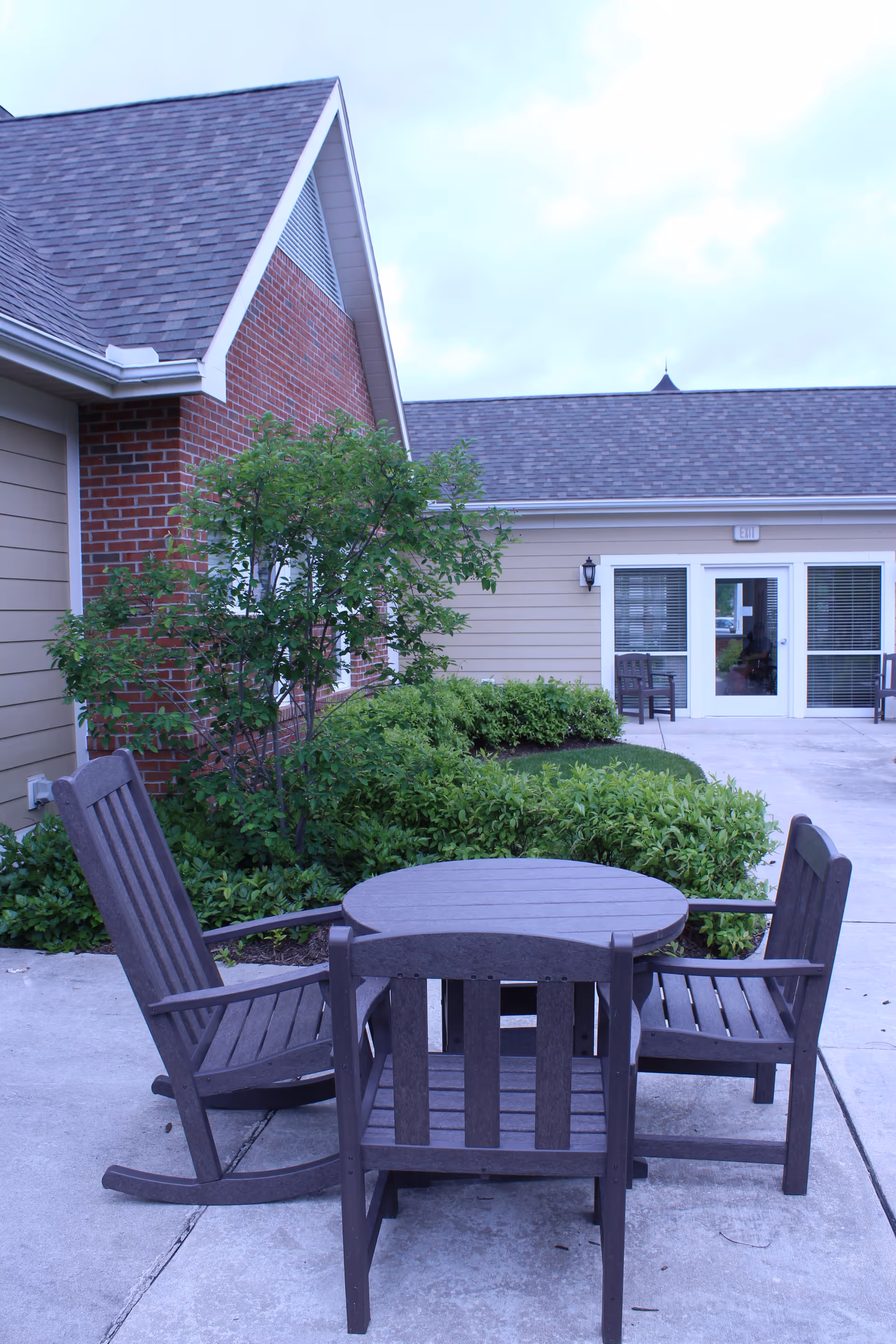Patio with a round wooden table and three chairs in front of a brick-and-siding building entrance and landscaping.