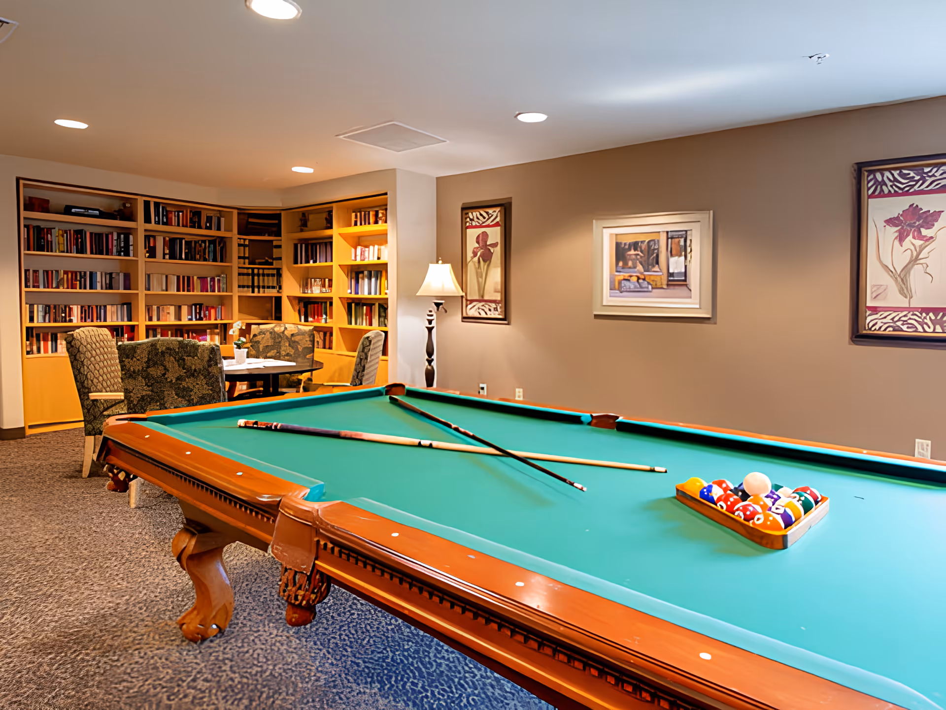A recreational room featuring a green pool table with two pool cues resting on it and a rack of billiard balls. In the background, there is a cozy seating area with patterned chairs and a small table, along with built-in wooden bookshelves filled with books. The walls are decorated with framed artwork and a standing lamp provides additional lighting.