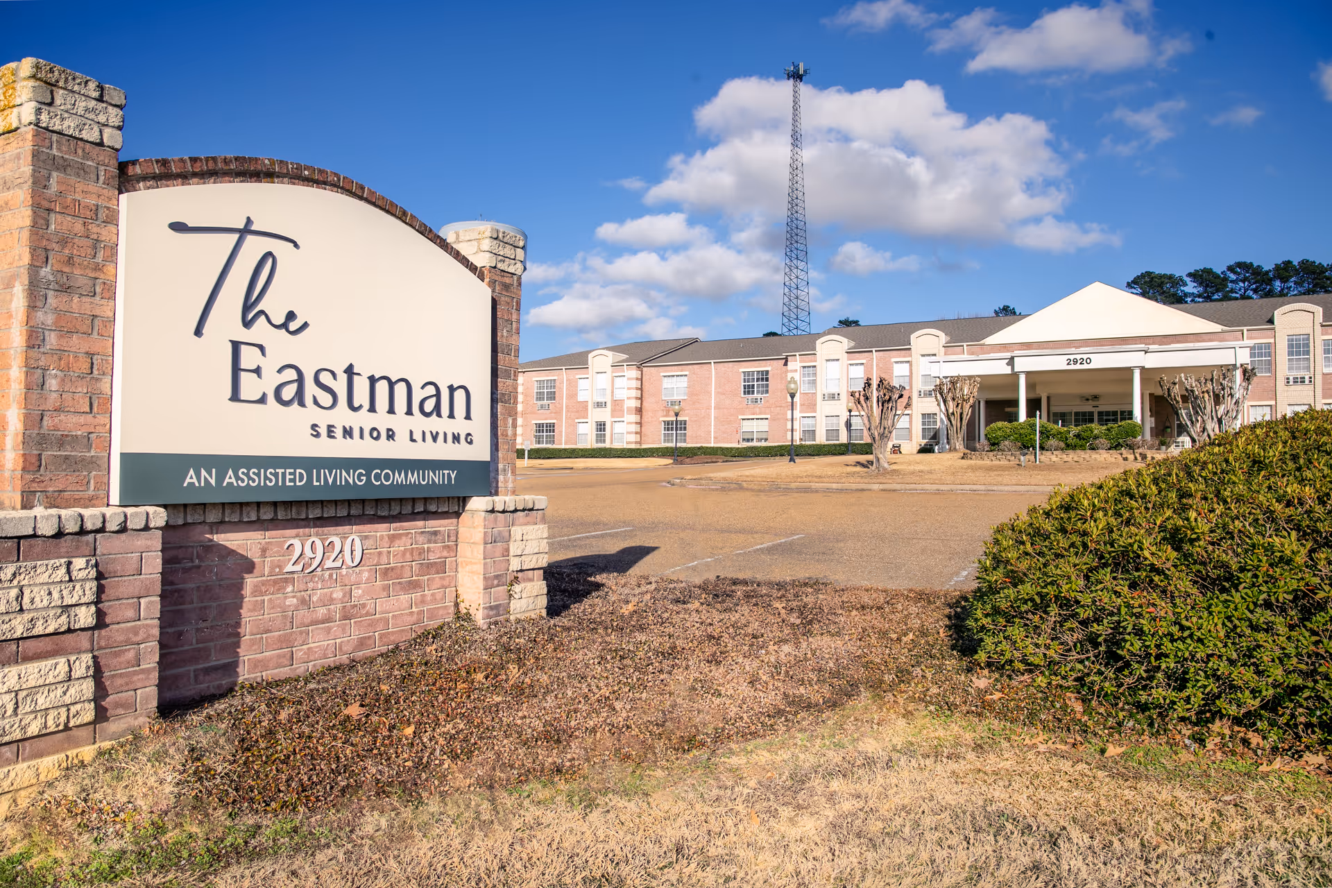 Exterior view of The Eastman Senior Living assisted living community building with a large sign in the foreground displaying the facility name and address 2920. The building is a two-story brick structure under a blue sky with some clouds.