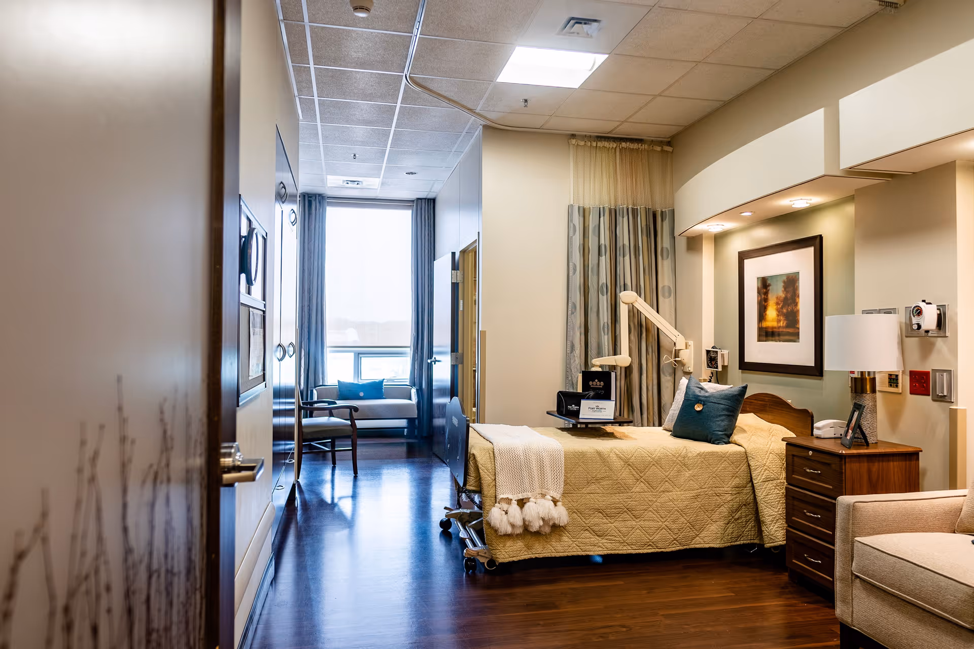 A well-lit, cozy bedroom in a care facility featuring a single bed with a yellow quilt and blue pillow, a bedside table with a lamp and phone, a framed picture on the wall, a beige armchair, and a window with curtains letting in natural light. The room has wooden flooring and a ceiling with recessed lighting.