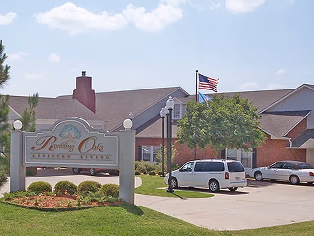Exterior view of a senior living facility with a sign that reads 'Rambling Oaks Assisted Living' in front of a brick building with a chimney, several parked cars, a tree, and an American flag flying on a flagpole.