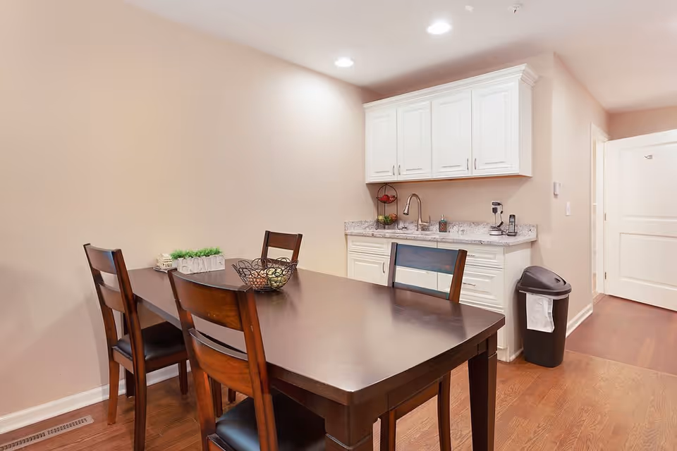 Dining area with a dark wood table and chairs beside a small white kitchenette with cabinets and sink.