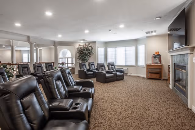 A spacious living room with multiple black leather recliner chairs arranged in rows facing a fireplace and a television mounted above it. The room has large windows with white blinds, a patterned carpet, and a popcorn machine on a wooden stand near the fireplace. There are also decorative plants and columns along the left side of the room.