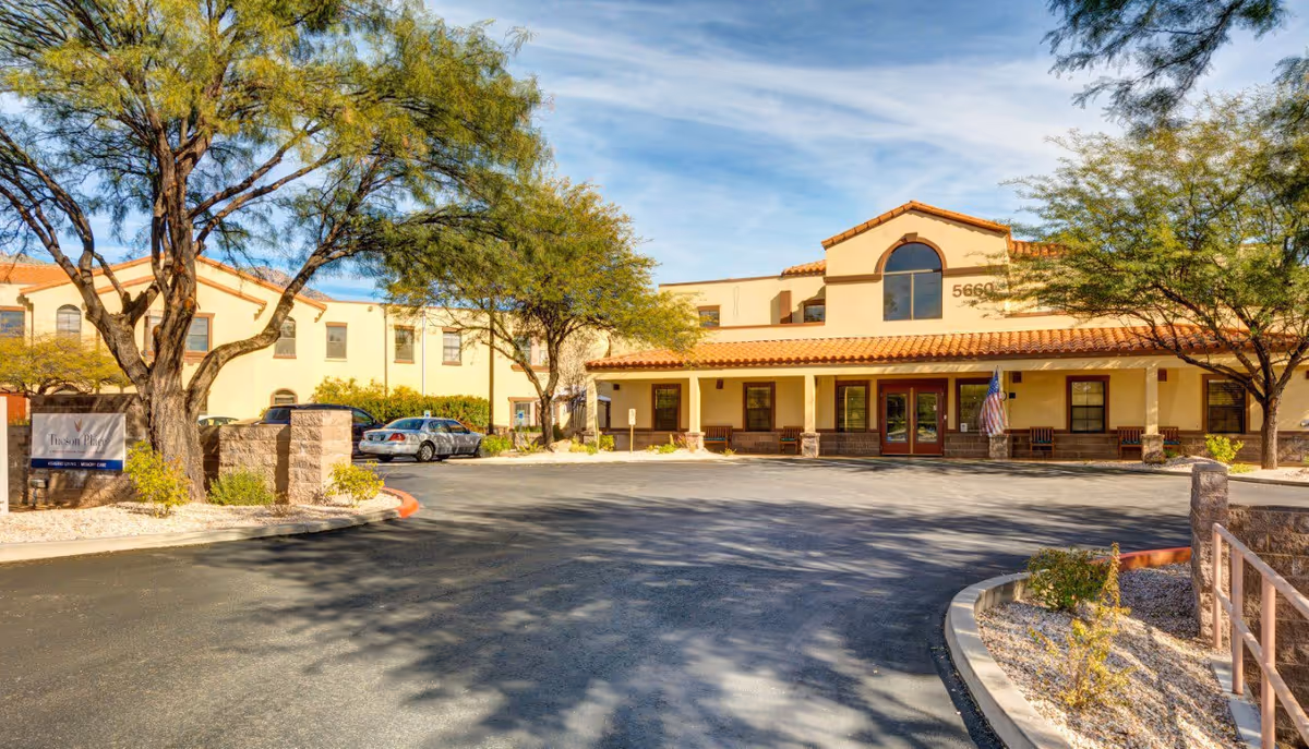 Exterior view of Tucson Place at Ventana Canyon, showing a two-story building with a tiled roof, arched windows, and a driveway in front. Trees and desert landscaping surround the building under a partly cloudy sky.