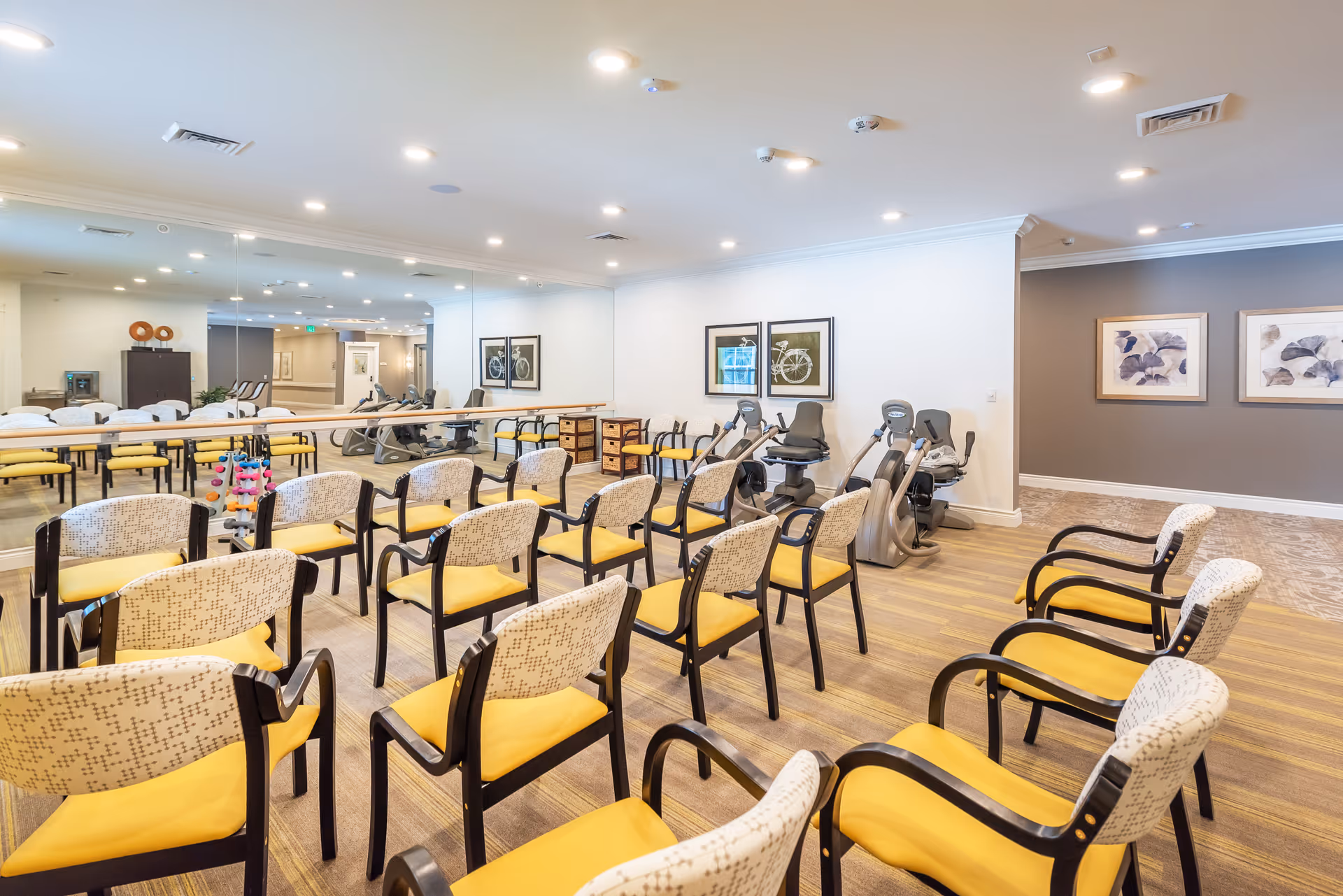 A bright and spacious room with multiple rows of chairs featuring yellow cushions and patterned backs, arranged facing a mirrored wall. Behind the chairs, there are exercise machines including stationary bikes and a recumbent bike. The room has light-colored walls with framed artwork and a wooden floor.
