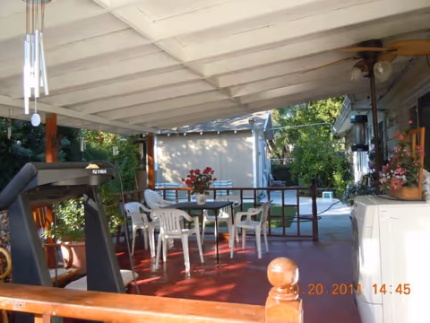 Covered outdoor patio area with a red floor, white plastic chairs and tables, a treadmill, potted plants, and a ceiling fan. The patio overlooks a garden with trees and a small shed in the background.