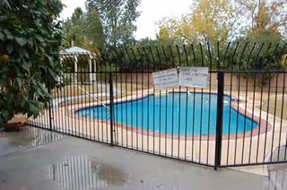 Outdoor fenced pool area with blue water, concrete deck, and trees in the background.