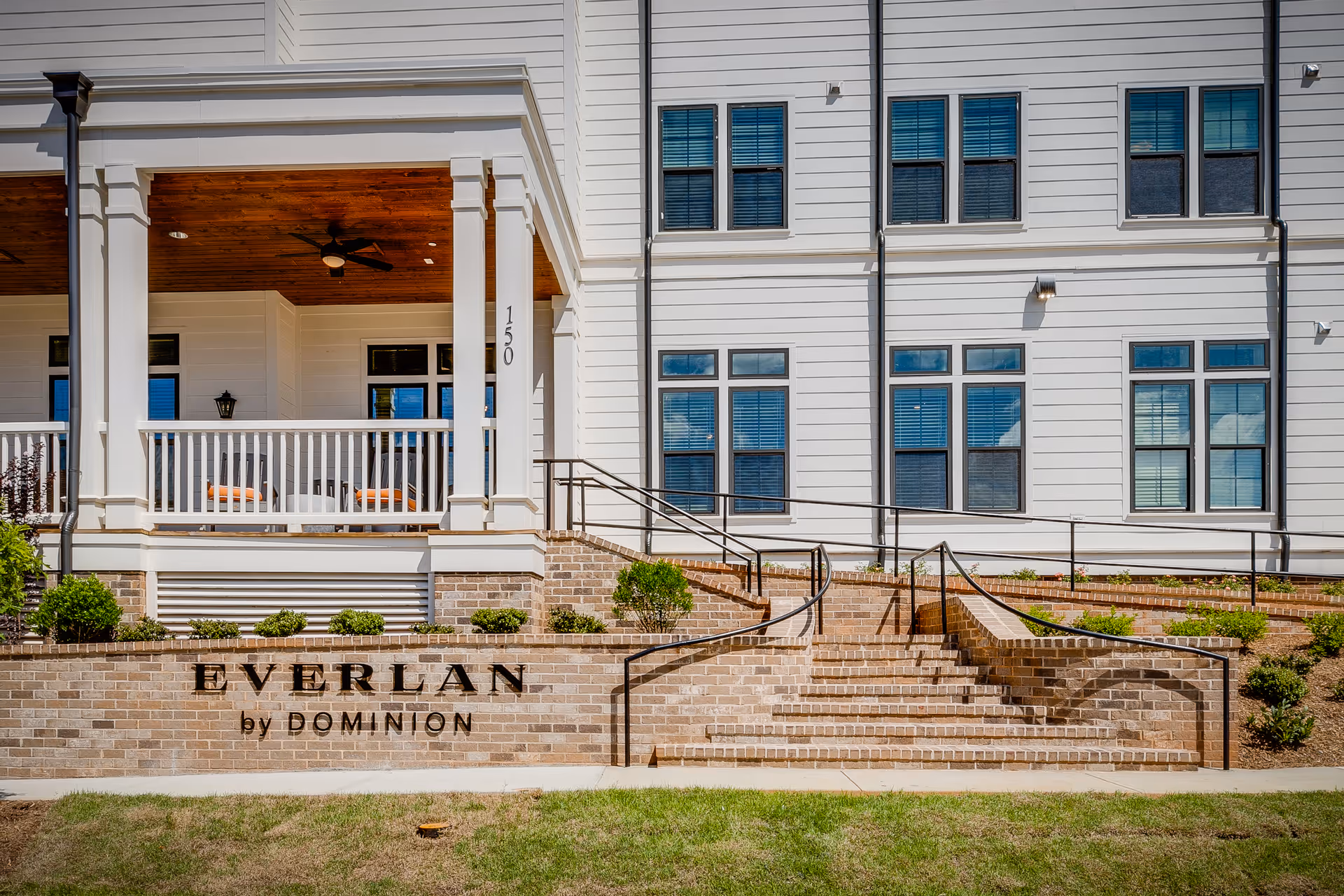 Front exterior view of Everlan by Dominion facility showing a white building with multiple windows, a covered porch with ceiling fan, brick stairs with black handrails, and landscaping with small bushes.