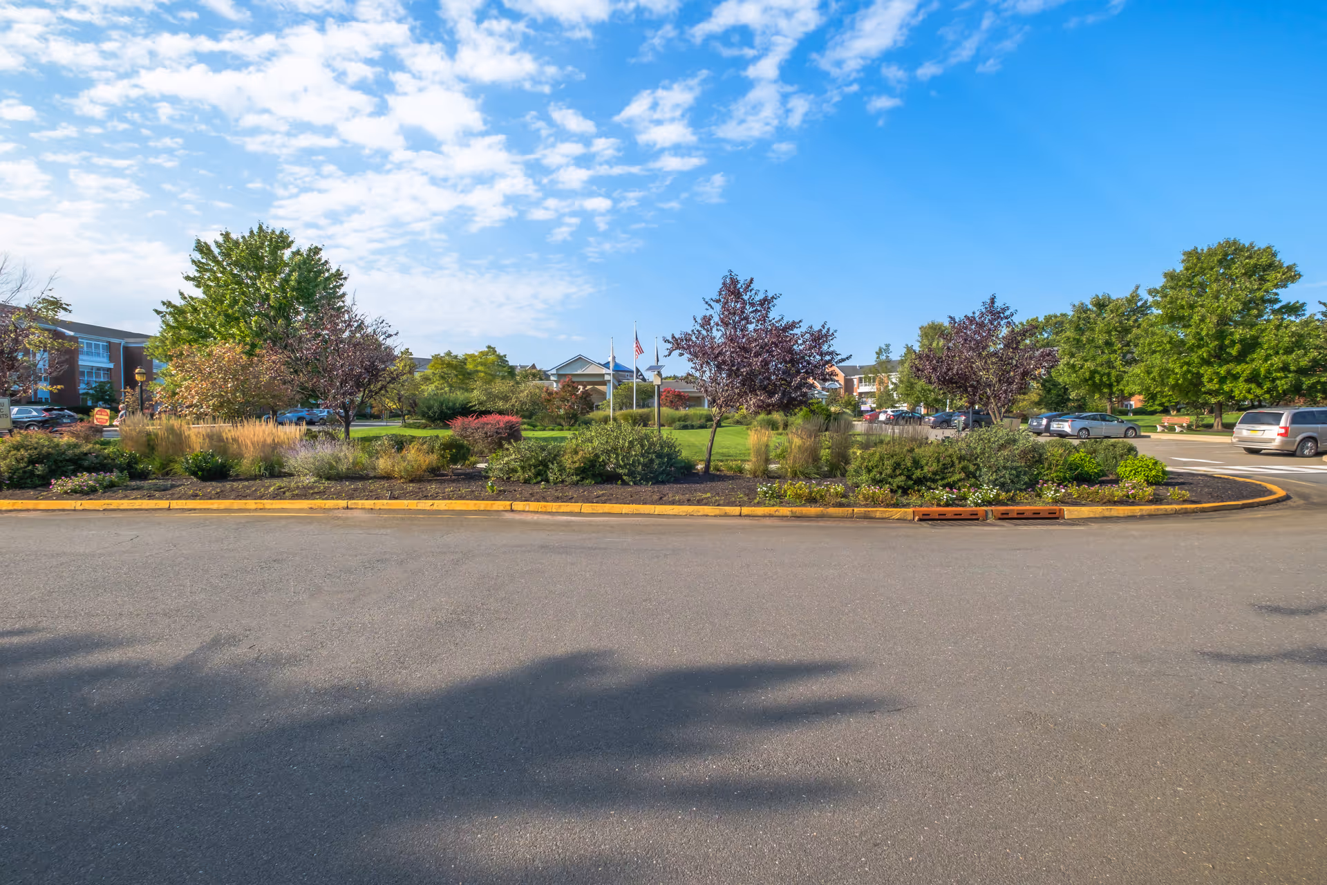View of a landscaped roundabout with various trees and shrubs in front of a senior living facility under a partly cloudy blue sky. Several cars are parked along the driveway surrounding the roundabout.