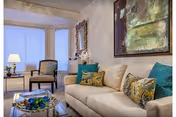 Bright living room with a beige sofa, patterned throw pillows, a glass coffee table, accent chairs, a lamp, and large bay windows.