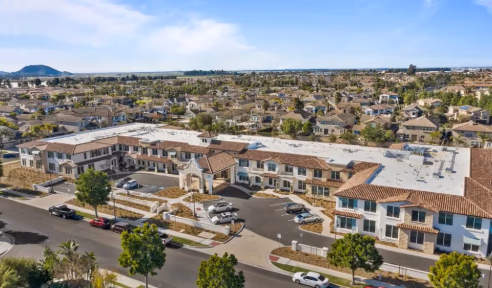 Aerial view of a large tiled-roof senior living building with a porte-cochère, parking lot and surrounding residential neighborhood.
