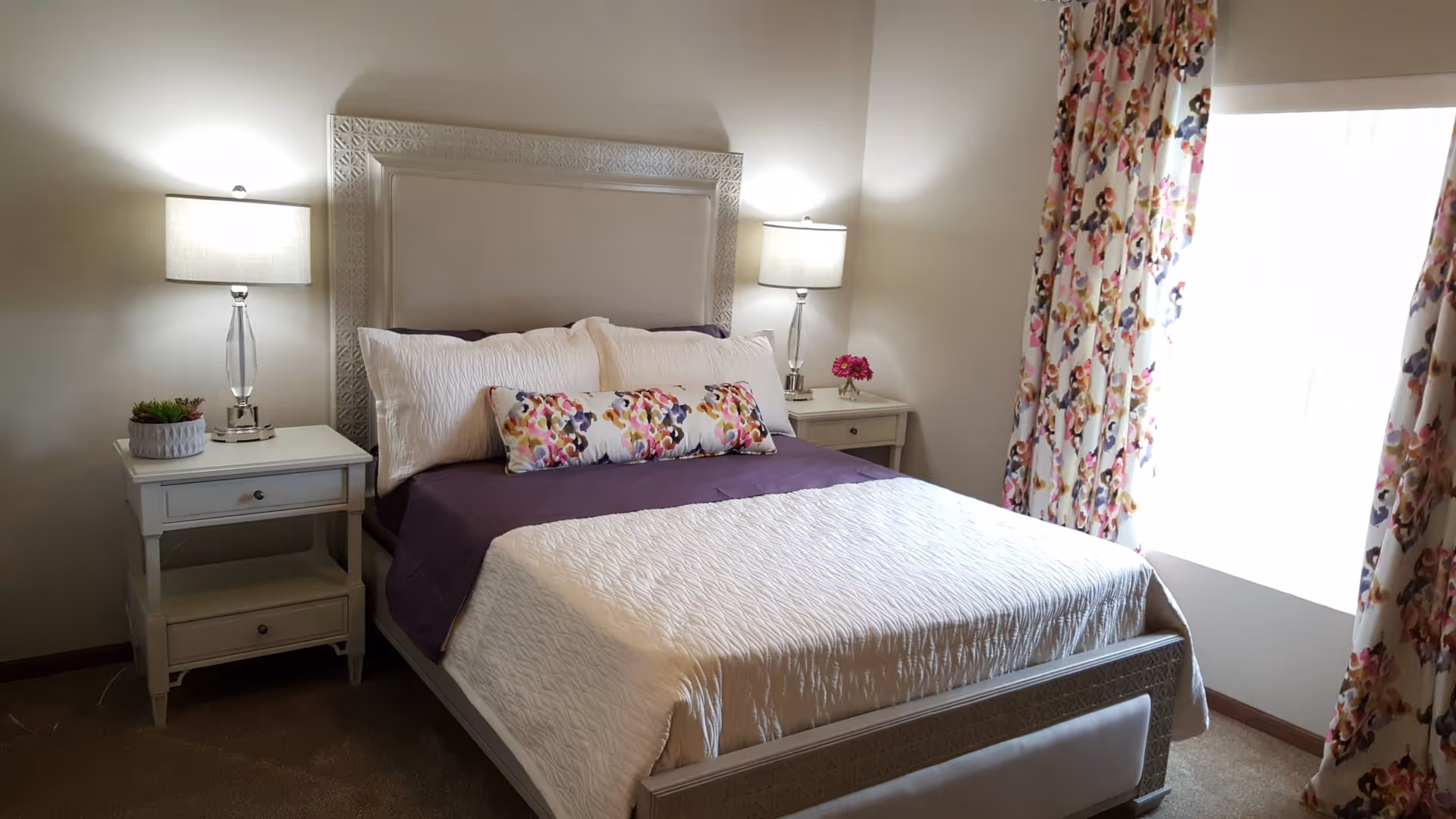 A neatly made bed with a white quilt and purple blanket in a bedroom. The bed has a white headboard with intricate designs and is flanked by two white nightstands, each with a lamp. There are floral-patterned curtains on the window to the right, and decorative pillows with a colorful floral pattern on the bed.