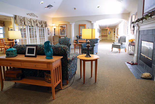A cozy senior living facility common area with patterned armchairs, a wooden side table with a lamp, a fireplace on the right, and a hallway leading to other rooms. The space is warmly lit and decorated with framed artwork and plants.