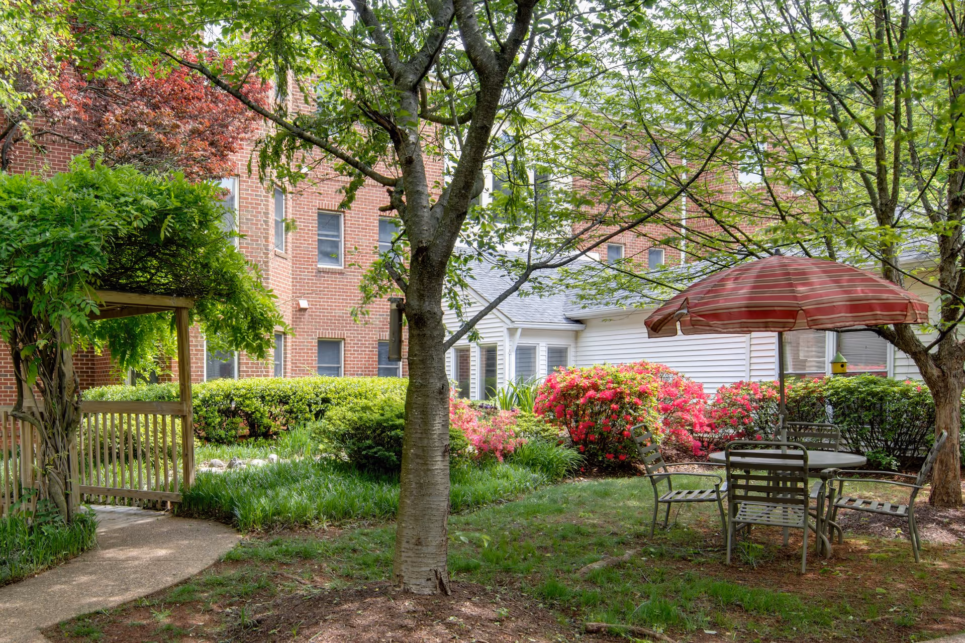 Outdoor garden area at The Manor House with green trees, bushes, and blooming flowers. There is a round table with four metal chairs and a red-striped umbrella providing shade. A wooden pergola covered with greenery is visible on the left side, and the background shows a brick building and a white siding structure.