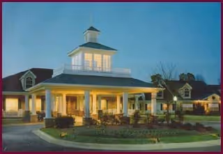 Front entrance of a well-lit senior living building with a covered porte-cochere, cupola, and landscaped circular driveway at dusk.