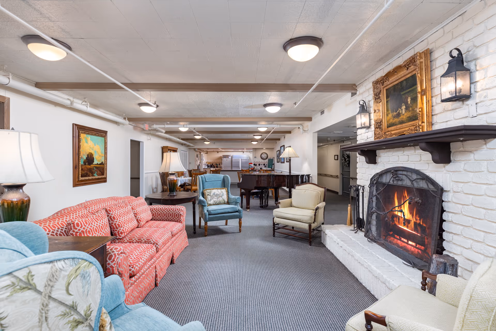 A cozy living room area in a retirement residence featuring a red patterned sofa, two blue armchairs, and a beige armchair arranged around a white brick fireplace with a fire burning. Above the fireplace is a framed painting and two wall-mounted lantern-style lights. In the background, there is a grand piano, dining tables, and chairs, with ceiling lights and exposed pipes on the ceiling.