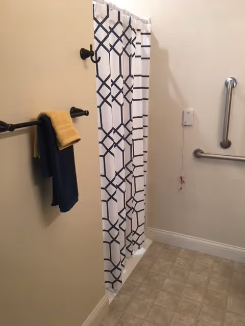 Bathroom with a shower area featuring a white shower curtain with a black geometric pattern. There are two towels hanging on a black towel rack on the left wall, one yellow and one dark blue. The walls are light-colored, and there are two silver grab bars mounted on the right wall near the shower. The floor has beige tiles.