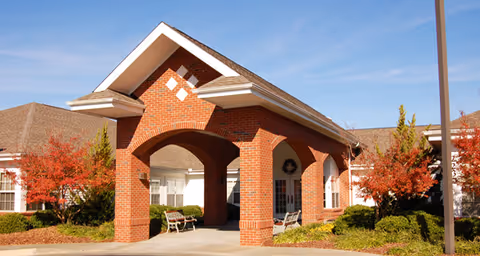 Entrance of a senior living facility with a large brick archway and peaked roof. There are benches under the archway and autumn-colored trees and shrubs surrounding the building. The sky is clear and blue.