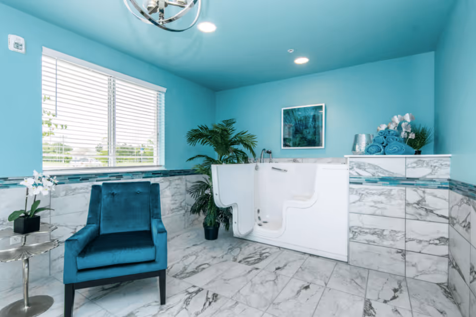 A bright bathroom with turquoise walls and ceiling, featuring a walk-in bathtub with a door, a blue upholstered armchair, a small side table with a white orchid plant, rolled turquoise towels on a marble ledge, a potted green plant, and a framed artwork on the wall.