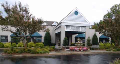 Exterior view of a single-story building with a covered entrance featuring a white lattice design and a small circular window near the roof peak. There are two benches and a multi-tiered water fountain surrounded by pink flowers in front of the entrance. The building is surrounded by well-maintained landscaping including bushes, trees, and green shrubs. The pavement in front is wet, indicating recent rain.