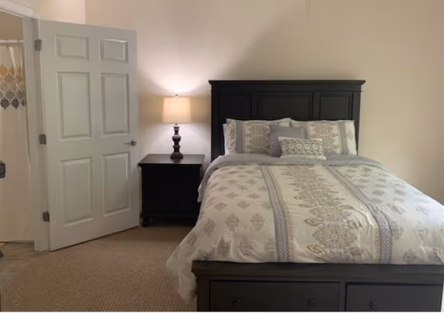 A bedroom with a neatly made bed featuring patterned bedding and multiple pillows. Next to the bed is a dark wooden nightstand with a lamp turned on. The door to an adjacent bathroom is open, revealing a shower curtain with a decorative pattern.