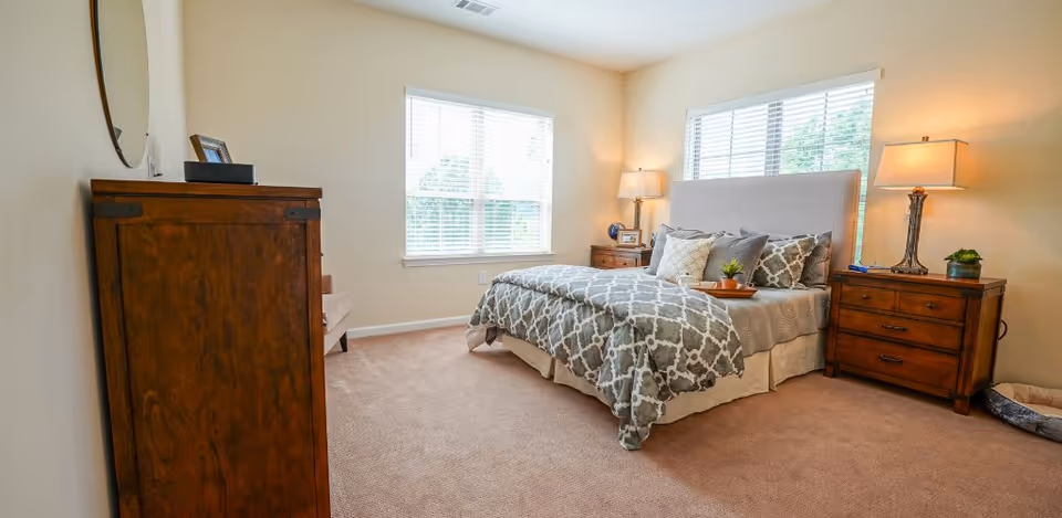Sunlit bedroom with a bed dressed in patterned bedding, wooden nightstands and a dresser beneath two windows.