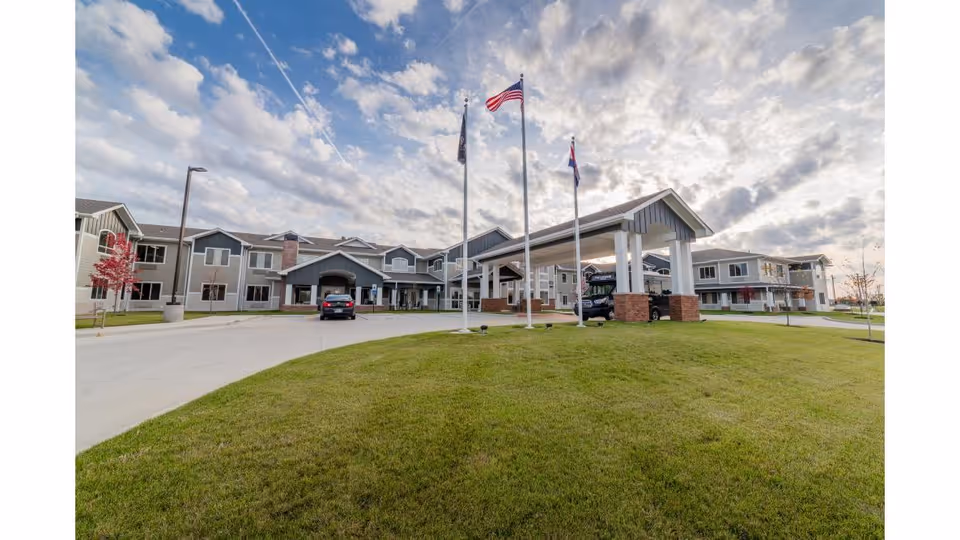 Exterior view of a senior living facility named The Landing of O'Fallon, showing a large two-story building with a covered entrance supported by brick pillars, three flagpoles with flags, a driveway, and a well-maintained grassy area under a partly cloudy sky.