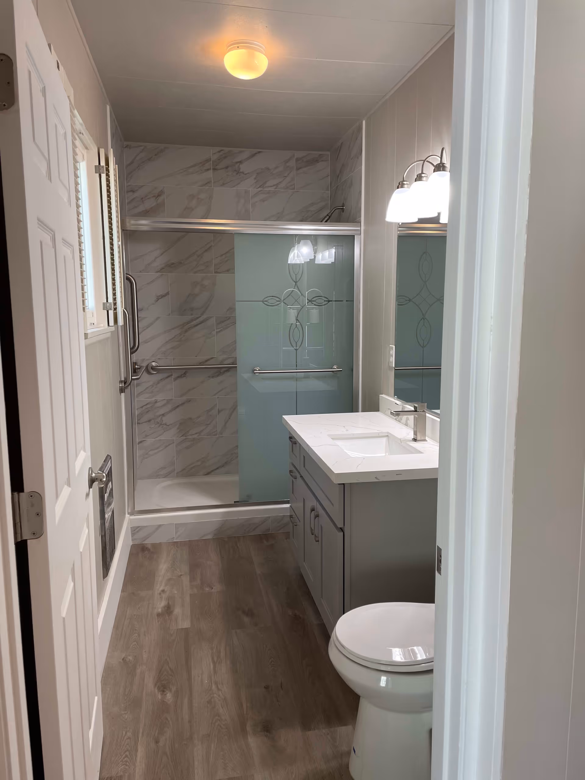 A modern bathroom with a white toilet, a gray vanity with a white countertop and sink, a large mirror with three light fixtures above it, and a shower with sliding glass doors and marble-patterned tiles. The floor is wood-style laminate, and the walls are light-colored.