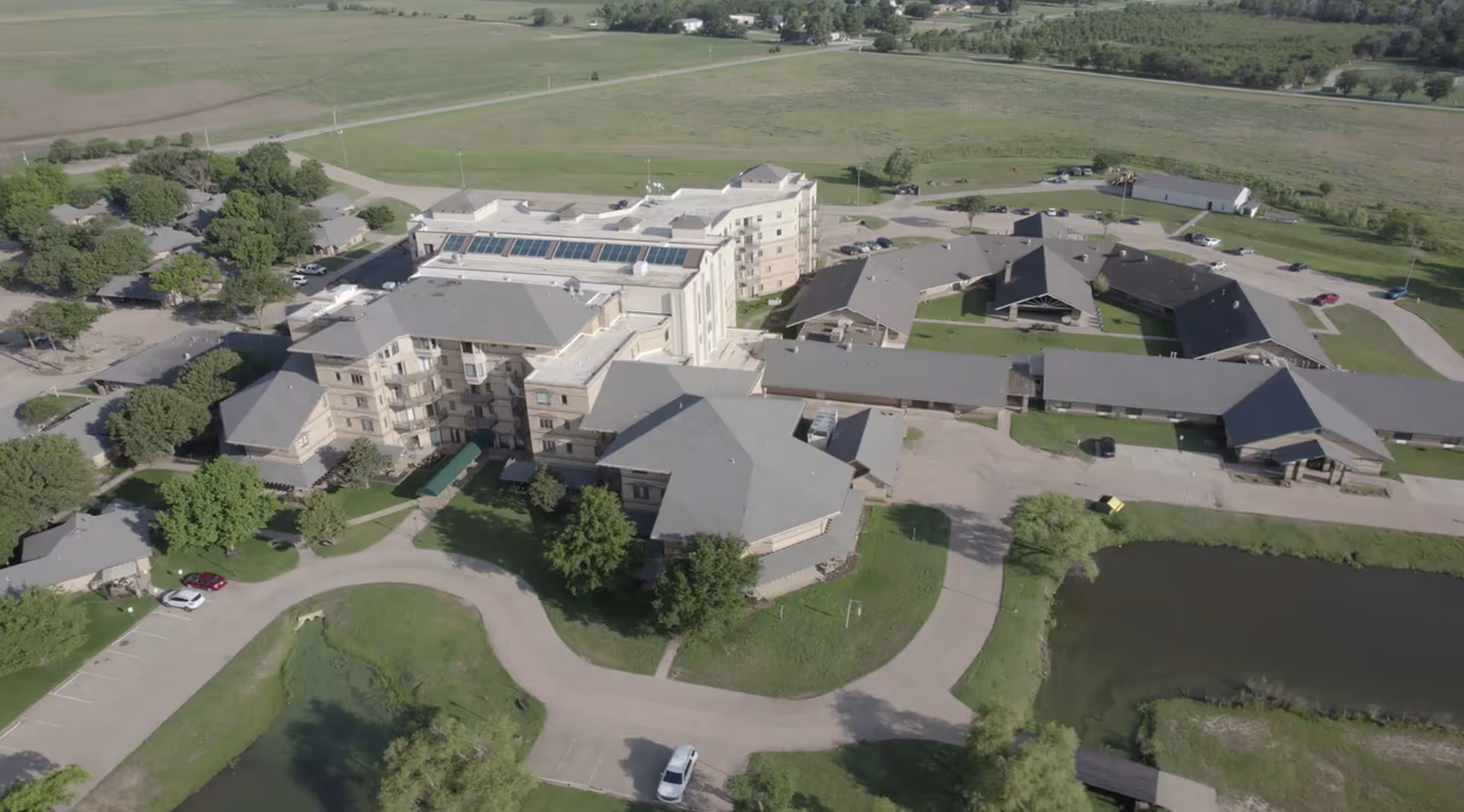 Aerial view of Baptist Village of Enid, showing multiple connected buildings with gray roofs surrounded by green lawns, trees, parking areas, and a pond. The facility is situated in a rural area with open fields in the background.