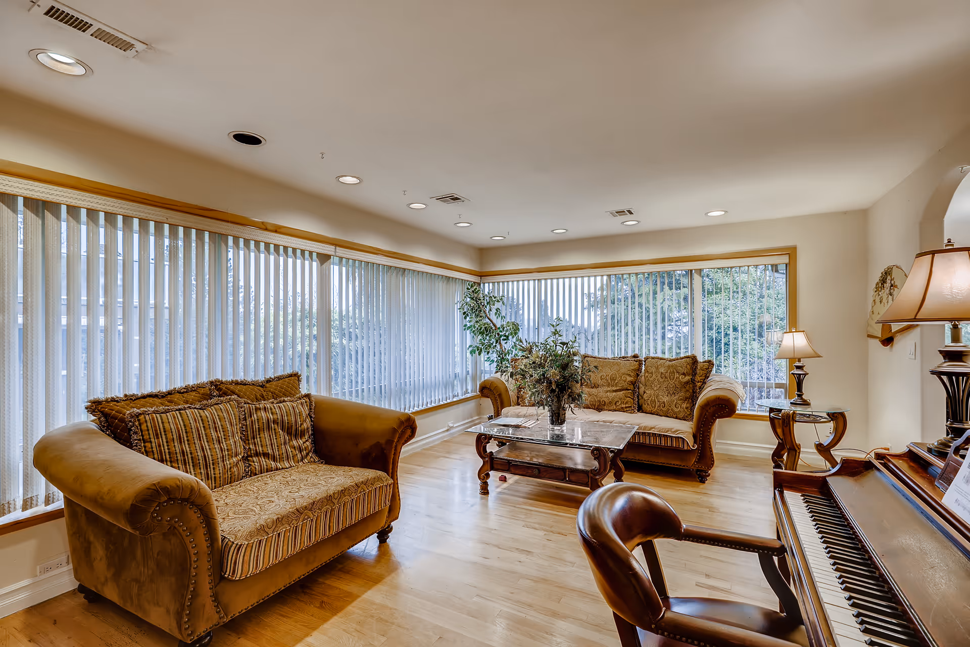 Spacious living room with upholstered sofas, a coffee table, piano, and large windows with vertical blinds.