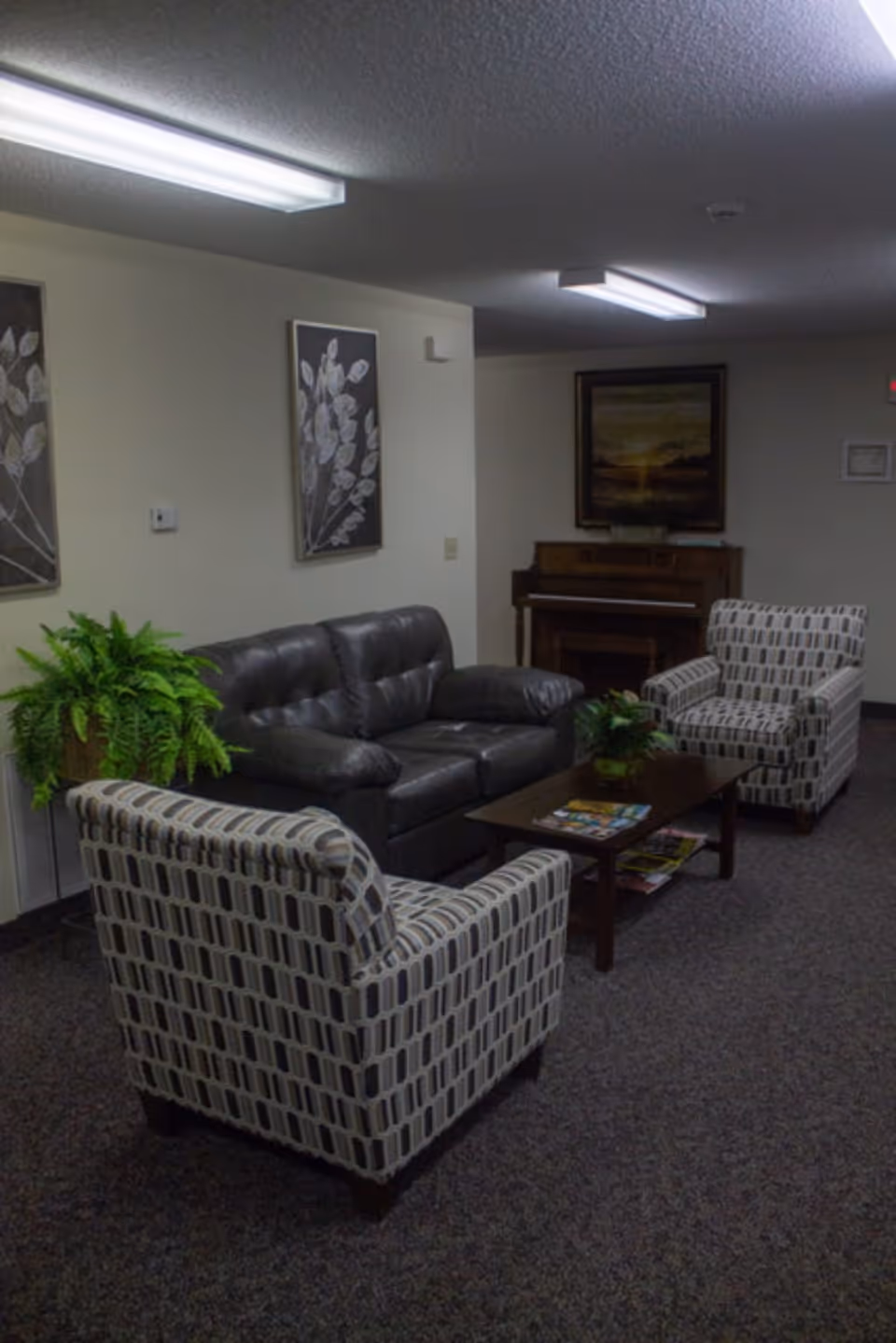 A cozy sitting area with a black leather couch, two patterned armchairs, a wooden coffee table with magazines, potted plants, and framed artwork on the walls. A piano is visible in the background under a framed painting.