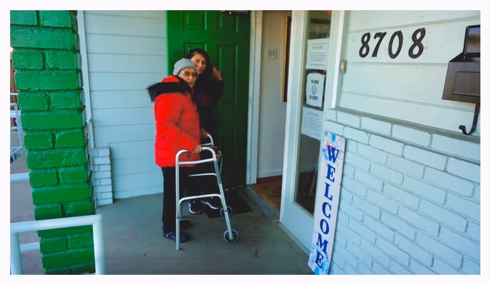 Two women standing outside the entrance of a building with the address number 8708. One woman is using a walker and wearing a red jacket and gray beanie, while the other woman stands behind her near a green door. A vertical welcome sign is leaning against the white brick wall next to the door.