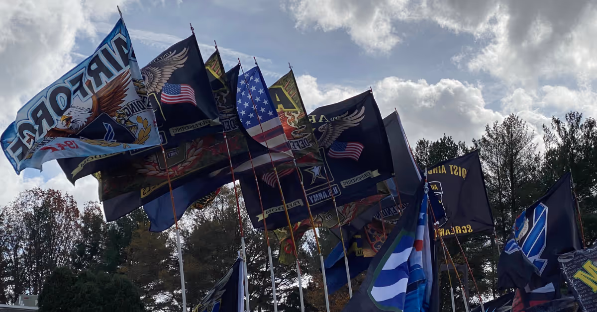 Multiple flags waving in the wind against a cloudy sky, including American flags and military-themed flags with eagles and various insignias, with trees in the background.